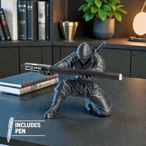 May include: A black ninja figurine holding a black pen with the text "TEST OF TIME". The figurine is kneeling and has a sword on its back. The image includes a pen.