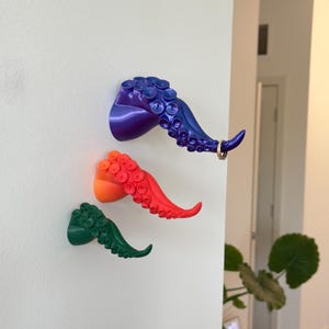 Puede incluir: Tres ganchos de pared con forma de tentáculos de pulpo en morado, naranja y verde. El tentáculo morado tiene un anillo dorado en el extremo. Cada tentáculo presenta detalles de ventosas. Estos ganchos decorativos añaden un toque náutico único.