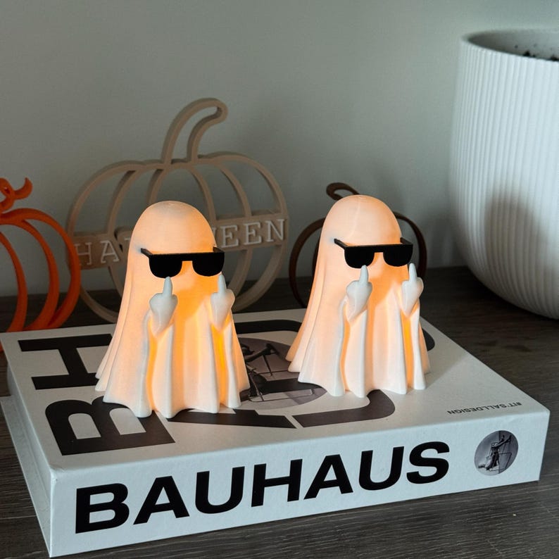 Puede incluir: Dos figuras de fantasmas iluminados con gafas de sol negras y el dedo coraz&oacute;n levantado. Los fantasmas est&aacute;n sobre un libro con la palabra "BAUHAUS". El fondo incluye decoraciones de calabazas y una maceta blanca.