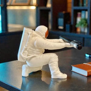 Puede incluir: Figura blanca de astronauta arrodillado, sosteniendo un bolígrafo negro. El astronauta tiene una visera naranja y una mochila blanca. La figura está sobre una superficie oscura, con un cuaderno marrón en el fondo.