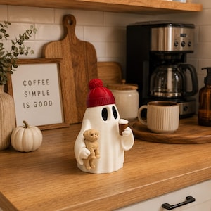 Könnte beinhalten: Eine weiße Geisterfigur mit einer roten Strickmütze mit Bommel. Der Geist hat schwarze, ovale Augen und hält einen kleinen beigen Welpen in einem Arm und einen braunen Kaffeebecher in der anderen Hand. Die Figur ist glatt und steht aufrecht.