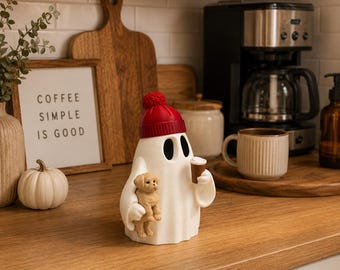 May include: A white ghost figurine wearing a red knit hat with a pom-pom. The ghost has black oval eyes and is holding a small tan puppy in one arm and a brown coffee cup in the other hand. The figurine is smooth and stands in an upright position.