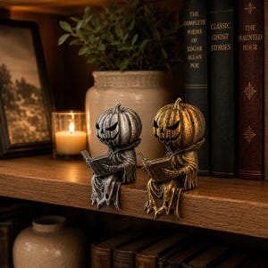 May include: Two shelf-sitting figurines featuring pumpkin-headed characters in hooded robes, each holding an open book. One figure has a metallic silver finish, while the other has a metallic gold finish. Both have carved, jagged facial features.