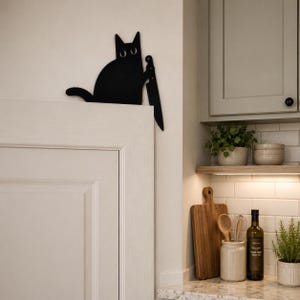 May include: A black metal cat silhouette wall decor piece featuring a cat holding a large kitchen knife. The cat has two circular cutouts for eyes and is positioned to hang over the top edge of a door or cabinet frame in a kitchen setting.