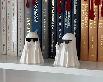 Puede incluir: Dos figuras de fantasmas blancos con gafas de sol y haciendo una peineta, colocadas en una estantería blanca frente a una fila de libros. La estantería también presenta gotas decorativas rojas. Los libros tienen varios títulos y colores.