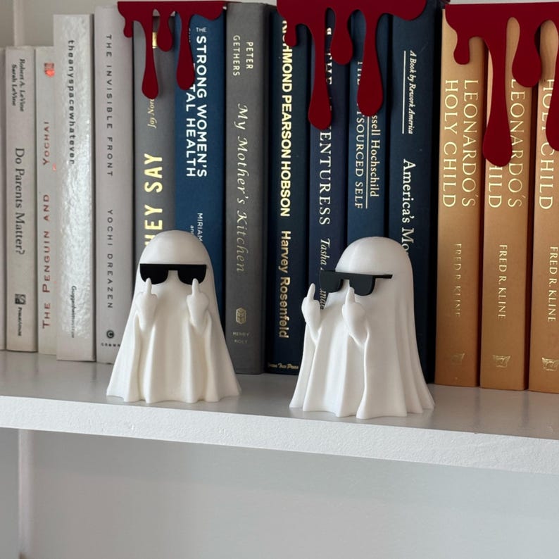 Puede incluir: Dos figuras de fantasmas blancos con gafas de sol y haciendo una peineta, colocadas en una estanter&iacute;a blanca frente a una fila de libros. La estanter&iacute;a tambi&eacute;n presenta gotas decorativas rojas. Los libros tienen varios t&iacute;tulos y colores.