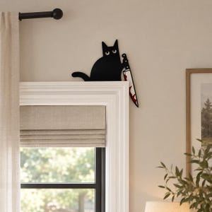 May include: A black cat wall decoration made of flat material, featuring a silhouette of a cat with wide eyes holding a large kitchen knife with red blood-like stains on the blade, positioned on the top corner of a white window frame.