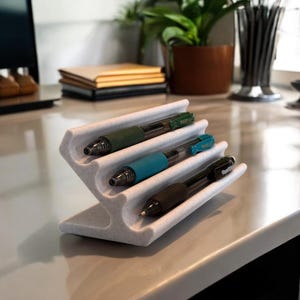 Pode incluir: Porta-canetas de plástico branco com quatro ranhuras para canetas. O porta-canetas tem uma superfície texturizada e está cheio de três canetas, uma verde, uma azul e uma preta.