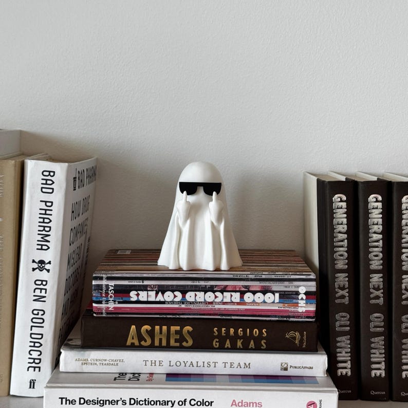 Puede incluir: Una figura de fantasma blanco con gafas de sol haciendo una peineta, colocada sobre una pila de libros. Los libros tienen t&iacute;tulos como "1000 Record Covers" y "Ashes". La figura a&ntilde;ade un toque de humor a una estanter&iacute;a.