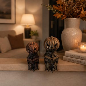May include: Two bronze-colored pumpkin-headed figures with carved, menacing facial expressions, each wearing a draped robe and holding an open book. These decorative figurines are positioned on a ledge, showcasing a weathered, metallic finish.