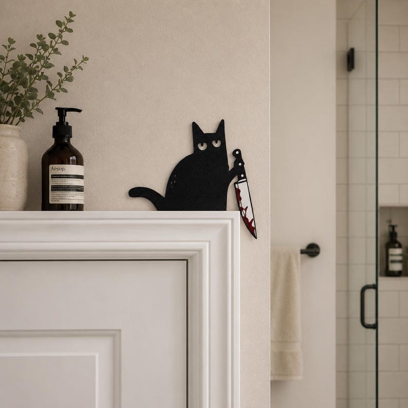 May include: A flat, black cat silhouette wall decoration featuring wide, white eyes and a long tail. The cat is holding a silver-colored knife with a red, blood-like stain on the blade. The item is positioned on the edge of a white door frame.