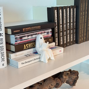 Puede incluir: Una figura de fantasma blanco de cer&aacute;mica leyendo un libro, colocada en un estante con una pila de libros. Los libros tienen t&iacute;tulos como "NEGOTIAUCTIONS" y "ASHES". El estante es blanco.