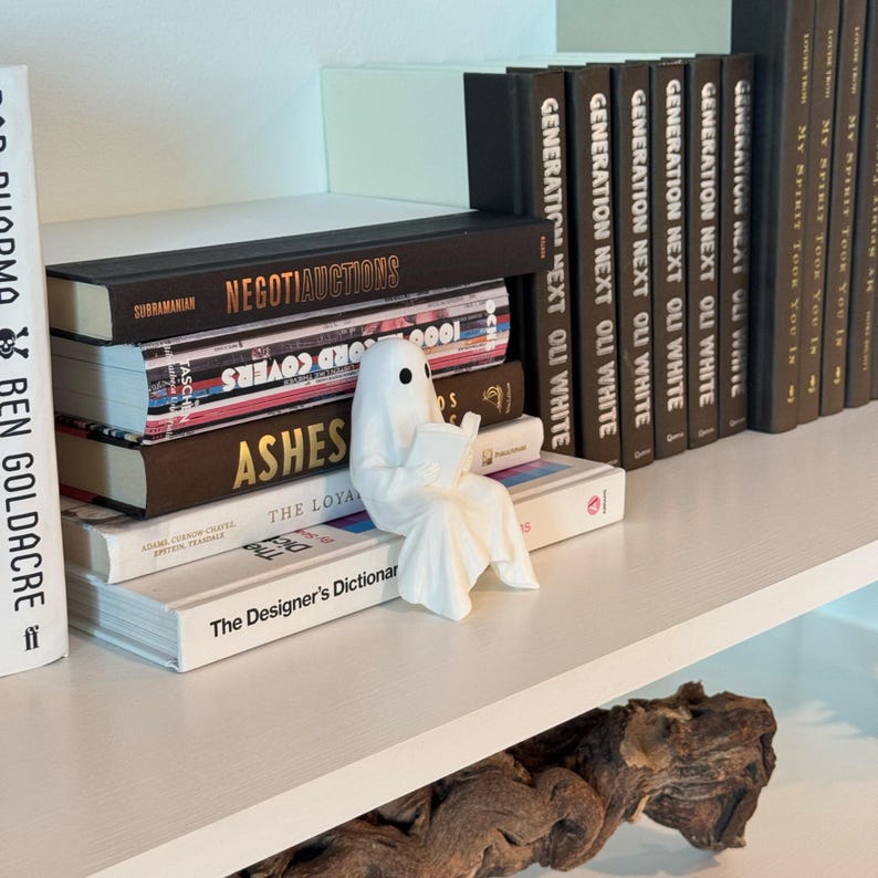 Puede incluir: Una figura de fantasma blanco de cer&aacute;mica leyendo un libro, colocada en un estante con una pila de libros. Los libros tienen t&iacute;tulos como "NEGOTIAUCTIONS" y "ASHES". El estante es blanco.