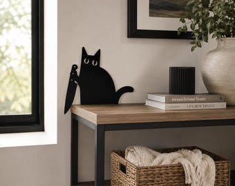 Pode incluir: Uma peça de arte de parede em metal preto plano com o formato de um gato segurando uma faca de cozinha grande. O gato tem uma silhueta simples com dois olhos circulares brancos e arregalados. A peça está posicionada verticalmente na parede.