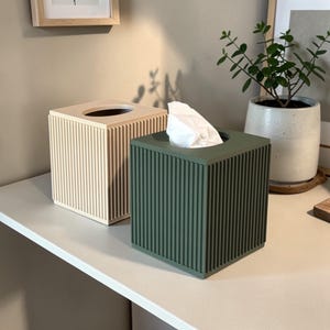 Puede incluir: Dos cubiertas de caja de pañuelos cuadradas. Una es de color beige claro y la otra verde oscuro. Ambas tienen un diseño de costillas verticales. La caja verde contiene pañuelos blancos. Decoración del hogar.