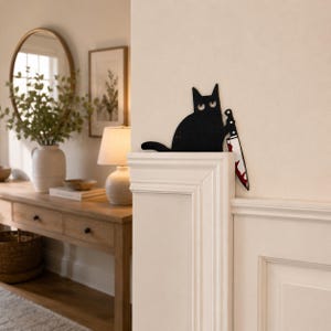 May include: A black metal wall art piece shaped like a cat with wide eyes, holding a silver knife with red blood-like stains on the blade. The cat figure is positioned to appear as if it is peeking over the top edge of a white door frame.