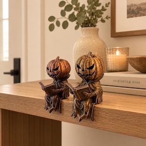 May include: Two bronze-coloured pumpkin-headed figures sitting on a ledge, each wearing a hooded robe and holding an open book. The figures feature carved jack-o'-lantern faces with sharp, triangular eyes and jagged mouths, finished with a weathered texture.
