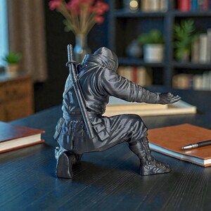 May include: A dark gray figurine of a ninja in a kneeling pose, with a katana strapped to their back. The ninja is wearing a hooded outfit and boots. The figurine is on a dark surface.