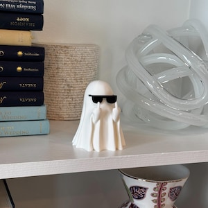 Puede incluir: Una figura de fantasma blanco con gafas de sol negras que hace un gesto obsceno. El fantasma est&aacute; sobre una estanter&iacute;a blanca con libros y objetos decorativos en el fondo.