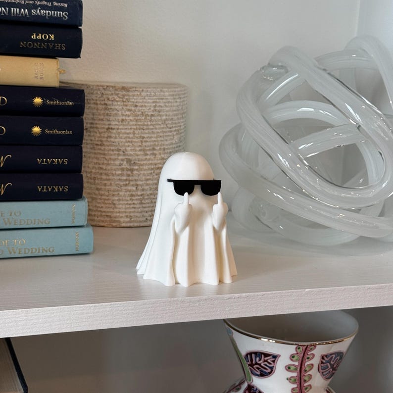 Puede incluir: Una figura de fantasma blanco con gafas de sol negras que hace un gesto obsceno. El fantasma est&aacute; sobre una estanter&iacute;a blanca con libros y objetos decorativos en el fondo.