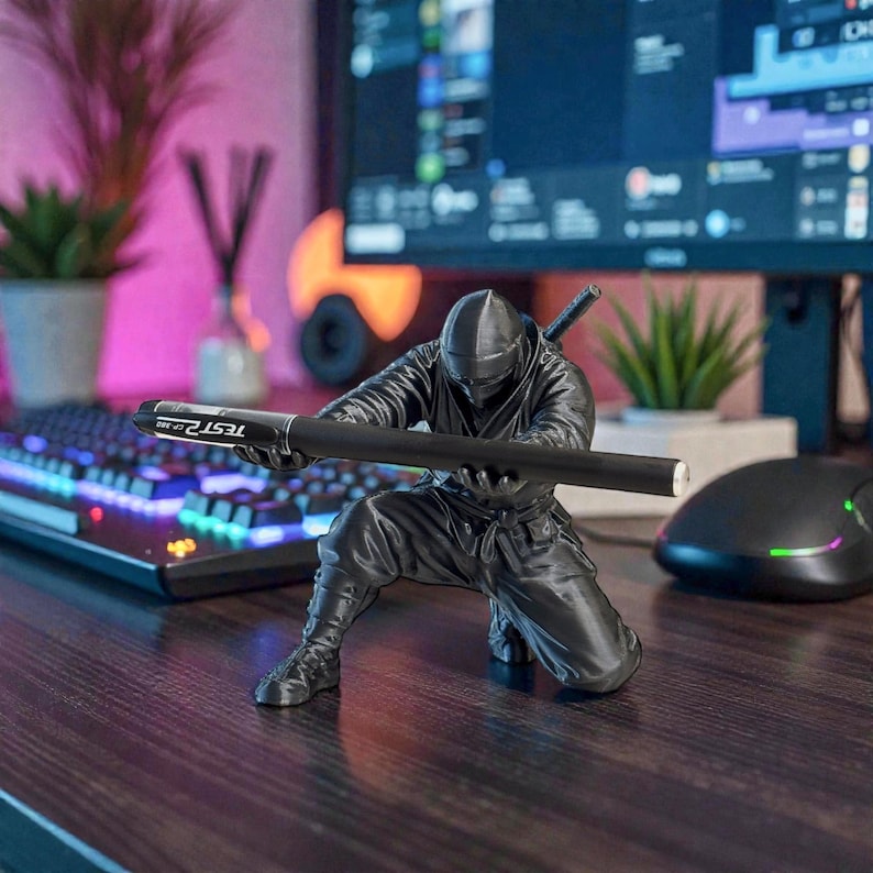 May include: A black ninja figurine holding a black pen with the text "TEST 2" on it. The ninja is in a crouching position, with a sword on its back. The background includes a keyboard, computer monitor, and a mouse.