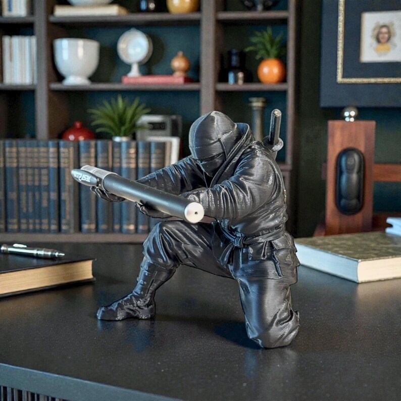 May include: A black ninja figurine in a crouching position, aiming a long, cylindrical object. The figure is detailed with a textured surface and is set against a backdrop of a bookshelf and desk.