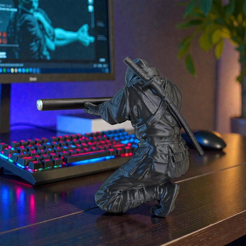 May include: A black ninja figurine in a crouching position, holding a black cylindrical object. The figurine has a katana on its back. A computer keyboard with red and blue lights is in the background.
