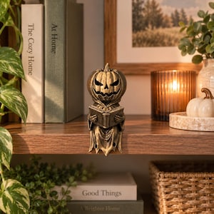 May include: A bronze-colored resin figurine of a pumpkin-headed creature wearing a hooded robe and holding an open book. The figure features a carved jack-o'-lantern face with a jagged grin and skeletal hands, designed to sit on a shelf edge.