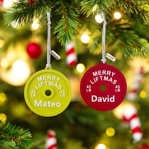 May include: Two Christmas tree decorations shaped like weight plates. One is lime green with "MERRY LIFTMAS", "45 LB", and "Mateo". The other is red with "MERRY LIFTMAS", "45 LB", and "David". Both are hanging on a Christmas tree.