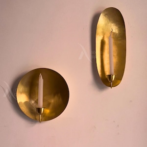 Puede incluir: Dos apliques de pared de latón, uno redondo y otro ovalado, cada uno con una vela blanca. Los apliques están montados en una pared rosa claro. Los candelabros tienen un diseño sencillo y elegante.