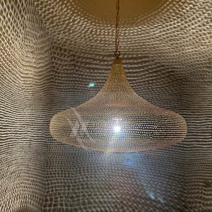 May include: A gold-colored, perforated metal pendant light fixture. The light is suspended from a chain and casts a patterned shadow on the surrounding wall. The fixture has a rounded, bell-like shape.