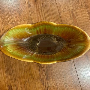 May include: An oval-shaped ceramic dish with a glossy finish. The dish features a blend of colors, including shades of green, brown, and gold. The interior has a dark brown center, transitioning to lighter hues towards the edges.