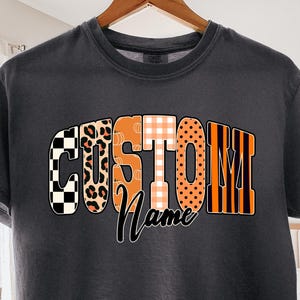 Peut inclure: Un t-shirt gris foncé arborant le mot CUSTOM en grandes lettres épaisses, chacune avec un motif unique incluant des damiers noirs et blancs, des taches de léopard, des carreaux orange et des pois, avec le mot Name écrit en cursif noir.