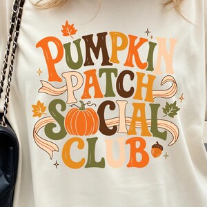 May include: An off-white short-sleeve t-shirt featuring a graphic print that reads PUMPKIN PATCH SOCIAL CLUB in orange, brown, and green lettering. The design includes a central orange pumpkin, autumn leaves, and an acorn illustration.