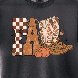May include: A charcoal gray t-shirt featuring a graphic print of the word FALL. The letter F has a brown and white checkerboard pattern, while the A, L, and L incorporate cowboy boots, pumpkins, acorns, and a lasso in warm earth tones.