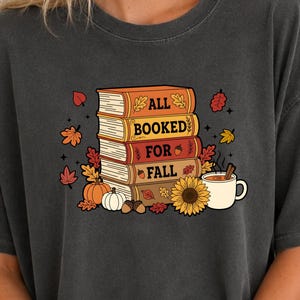 May include: A charcoal grey short-sleeve t-shirt featuring a graphic of a stack of four books with the text ALL BOOKED FOR FALL. The design includes colourful autumn leaves, a pumpkin, a white gourd, an acorn, a sunflower, and a mug of coffee.