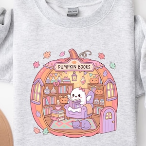 May include: Light gray sweatshirt featuring a whimsical illustration of a ghost reading inside a pumpkin-shaped library. The pumpkin has shelves filled with books and decorative items. The text "PUMPKIN BOOKS" is arched above the pumpkin. A cat is curled up on the floor.