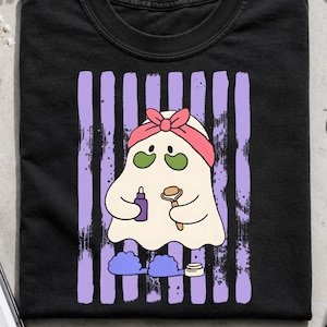 May include: A black t-shirt featuring a graphic of a white ghost wearing a pink headband and green under-eye patches. The ghost holds a purple serum bottle and a facial roller, with blue slippers at its feet, set against vertical purple stripes.