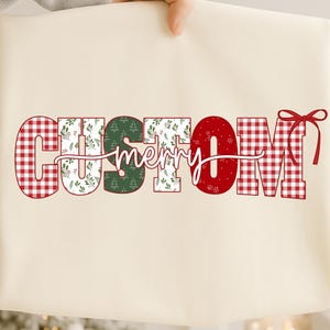 Puede incluir: Bolso tote color crema con la palabra "CUSTOM" en letras a cuadros rojos y estampado floral, y "merry" en cursiva. La letra "O" es roja con copos de nieve, y la letra "M" es a cuadros rojos. Un lazo rojo está a la derecha.