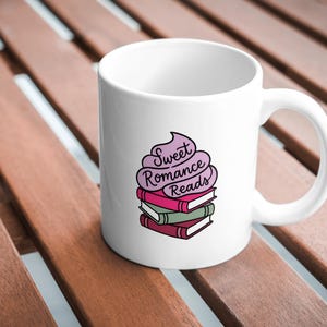 May include: White ceramic mug featuring a stack of books with the words "Sweet Romance Reads" in a whimsical font. The mug has a curved handle and is set against a wooden surface.
