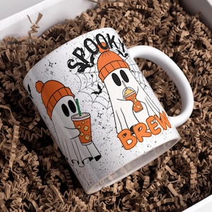 May include: White ceramic mug with a Halloween theme. The mug features two cartoon ghosts wearing orange beanies and holding iced drinks. The words "SPOOKY BREW" are printed on the mug. The mug is inside a white box filled with brown paper.