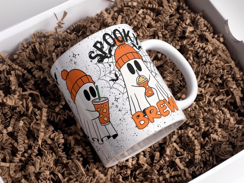 May include: White ceramic mug with a Halloween theme. The mug features two cartoon ghosts wearing orange beanies and holding iced drinks. The words "SPOOKY BREW" are printed on the mug. The mug is inside a white box filled with brown paper.