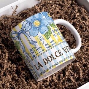 May include: White ceramic mug with a floral design featuring blue flowers and a yellow and white striped background. The mug has the text "LA DOLCE VITA" and "BUON APPETITO!" printed on it. The mug is inside a white box with brown paper filler.