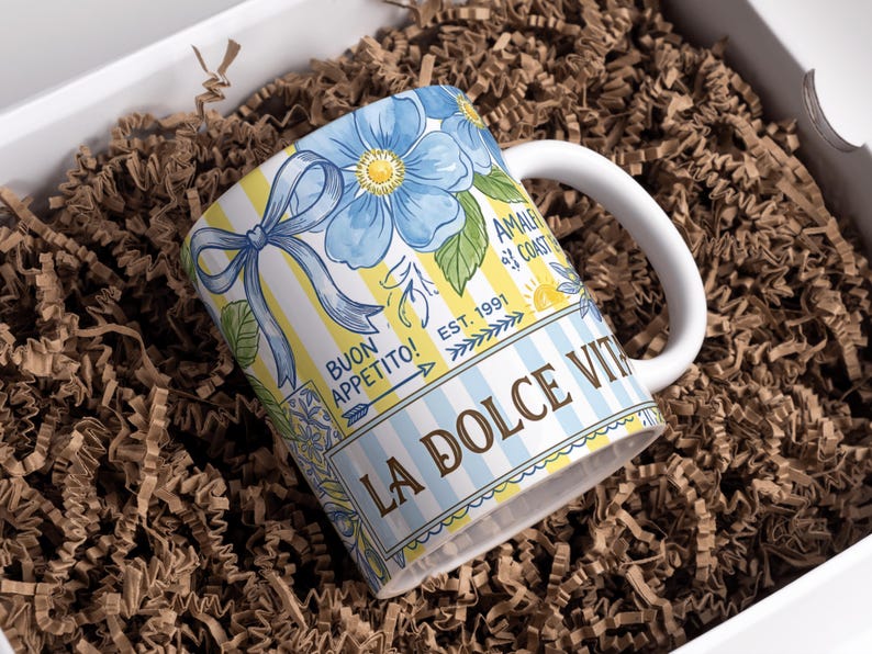 May include: White ceramic mug with a floral design featuring blue flowers and a yellow and white striped background. The mug has the text "LA DOLCE VITA" and "BUON APPETITO!" printed on it. The mug is inside a white box with brown paper filler.