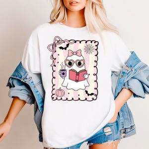 May include: White t-shirt featuring a cartoon ghost reading a book and holding a coffee cup. The ghost wears heart-shaped glasses and a bow. The design includes a pink pumpkin, bats, and a spiderweb, all within a decorative frame.