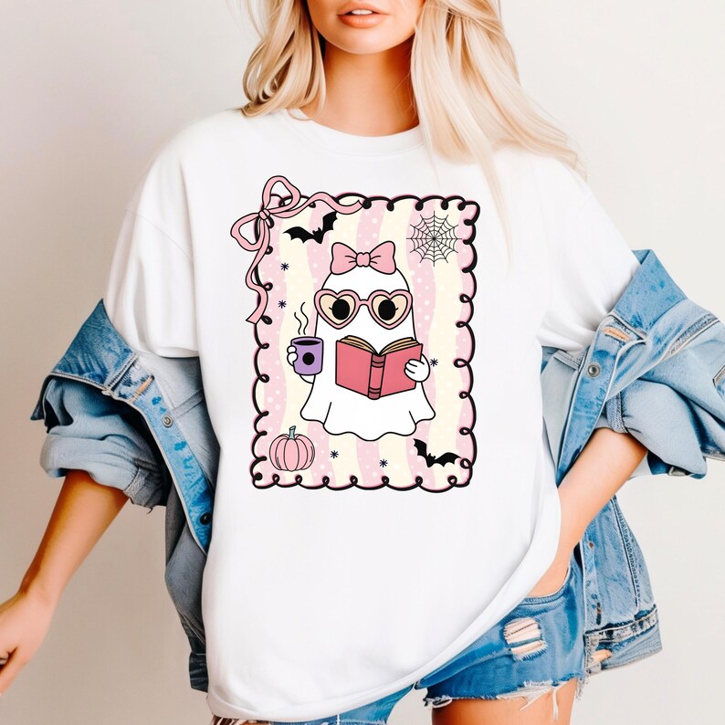 May include: White t-shirt featuring a cartoon ghost reading a book and holding a coffee cup. The ghost wears heart-shaped glasses and a bow. The design includes a pink pumpkin, bats, and a spiderweb, all within a decorative frame.