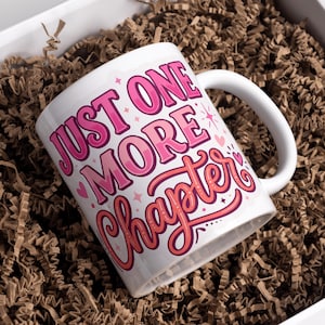 May include: White ceramic mug with the text "JUST ONE MORE Chapter" in pink and red lettering. The mug is inside a white box filled with brown crinkle paper.