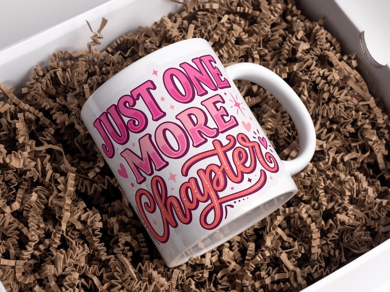 May include: White ceramic mug with the text "JUST ONE MORE Chapter" in pink and red lettering. The mug is inside a white box filled with brown crinkle paper.