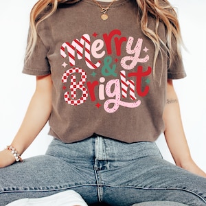 以下が含まれることがあります： 「Merry & Bright」の文字がデザインされた茶色のTシャツ。赤、ピンク、白の色使いで、「Bright」の文字にはチェック柄が施されています。カジュアルなスタイルのTシャツです。