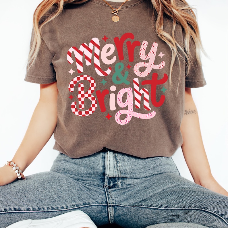 以下が含まれることがあります： 「Merry & Bright」の文字がデザインされた茶色のTシャツ。赤、ピンク、白の色使いで、「Bright」の文字にはチェック柄が施されています。カジュアルなスタイルのTシャツです。